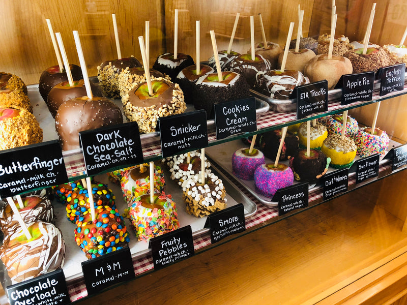 Display of various caramel apples with labels on a wooden shelf.